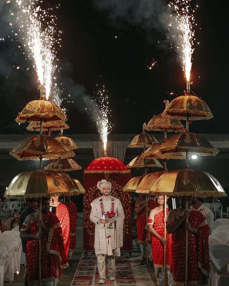 Groom making a grand Baraat entrance beneath ceremonial umbrellas and pyrotechnics